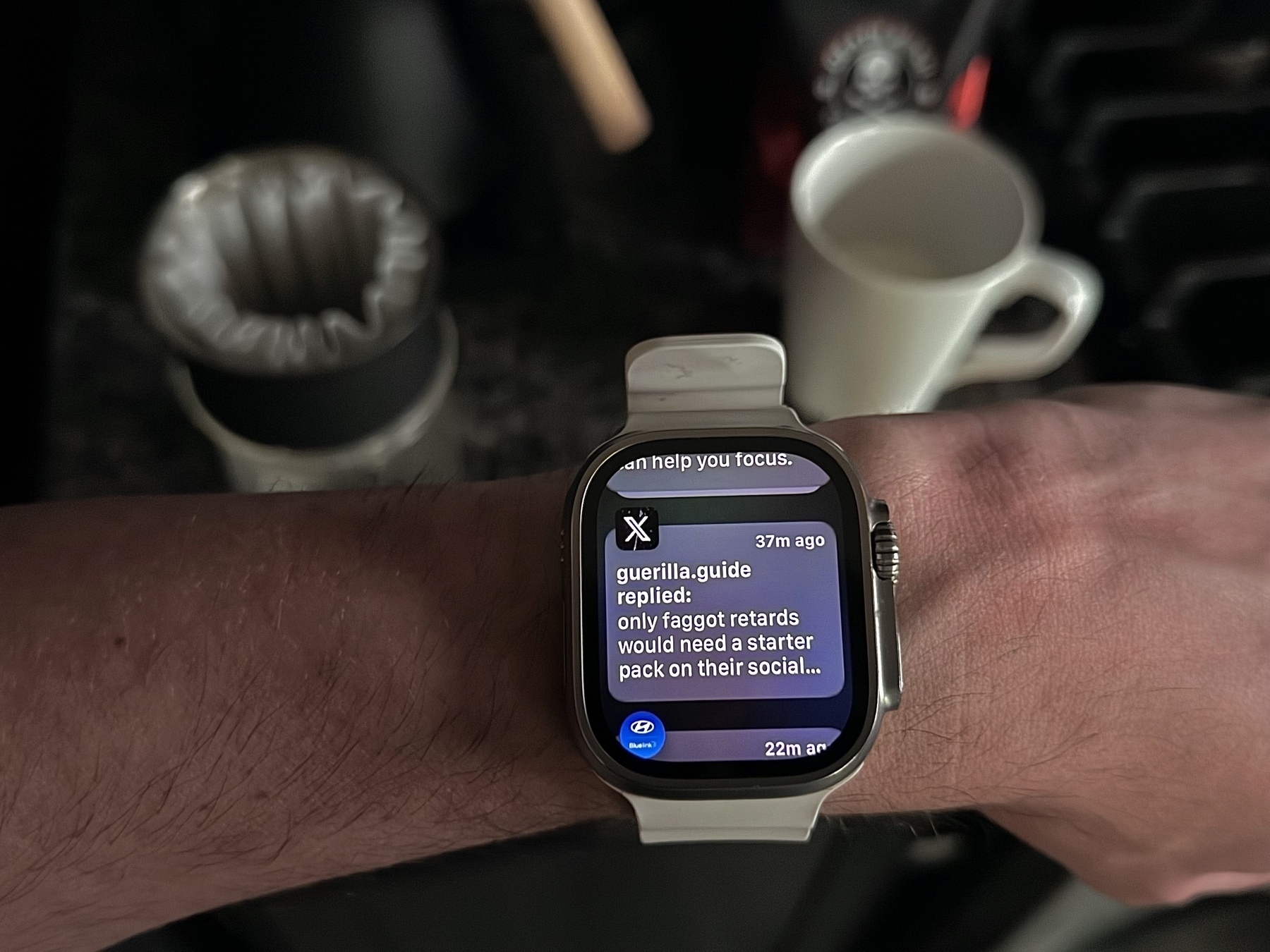 a photo of an Apple Watch on the user’s wrist and there’s a notification visible on it from X.com that is a reply from an anonymous user who is being verbally abusive